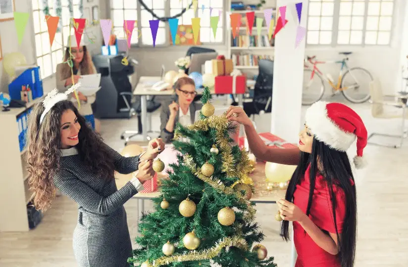 Two employees decorating christmas tree in office as they prepare for a party where the catering was booked in advance.